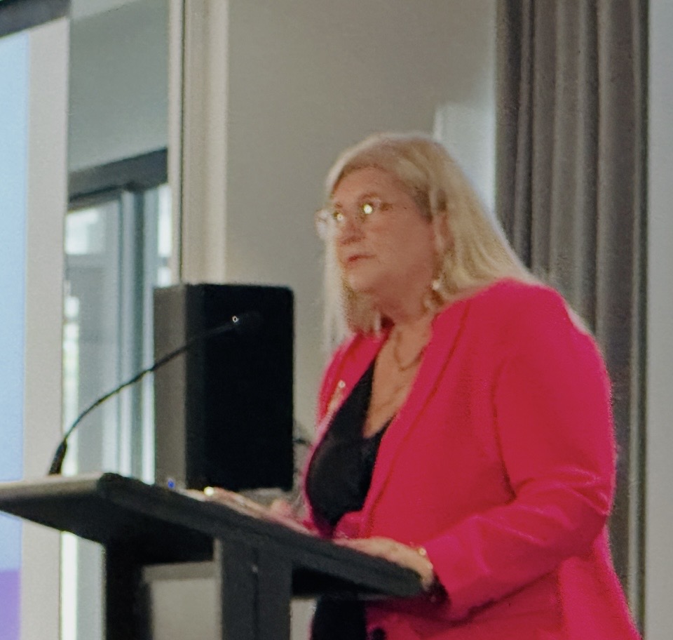 Dr Gabrielle Morrissey presenting from a lectern in a pink jacket